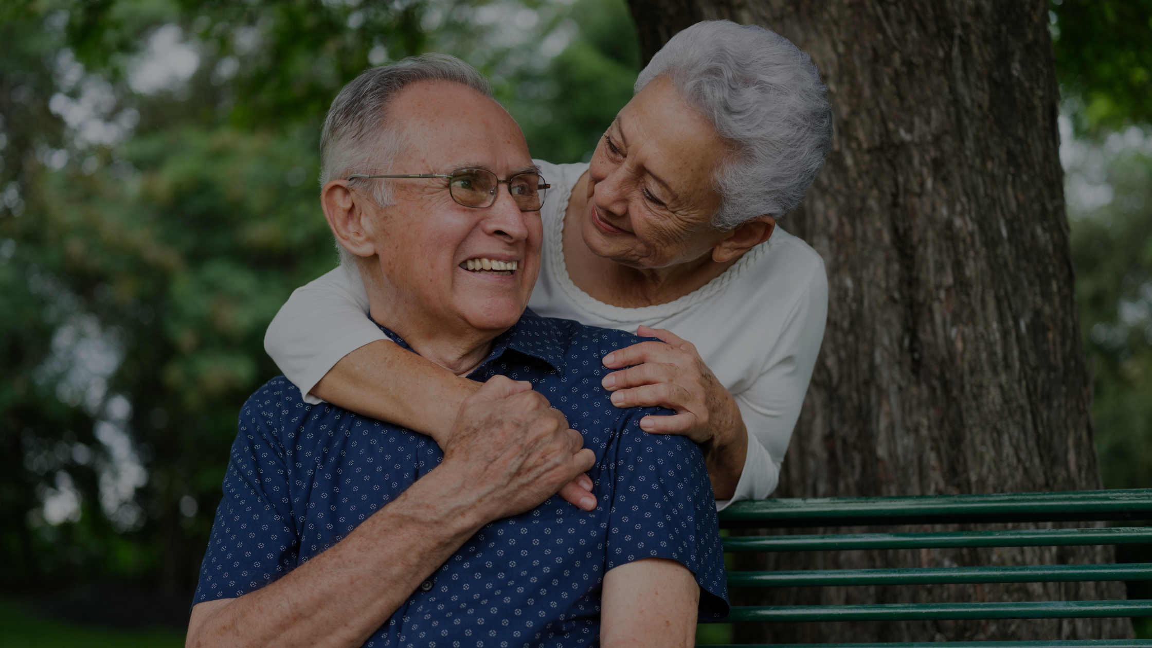 Two people share a joyful moment, sitting on a park bench under a tree, surrounded by greenery, smiling warmly at each other.