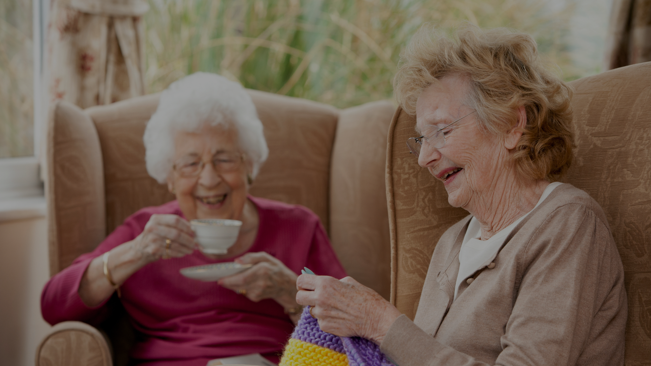 Two people sit in armchairs. One knits, the other drinks tea. Both are laughing, enjoying a light-hearted, cozy moment indoors.