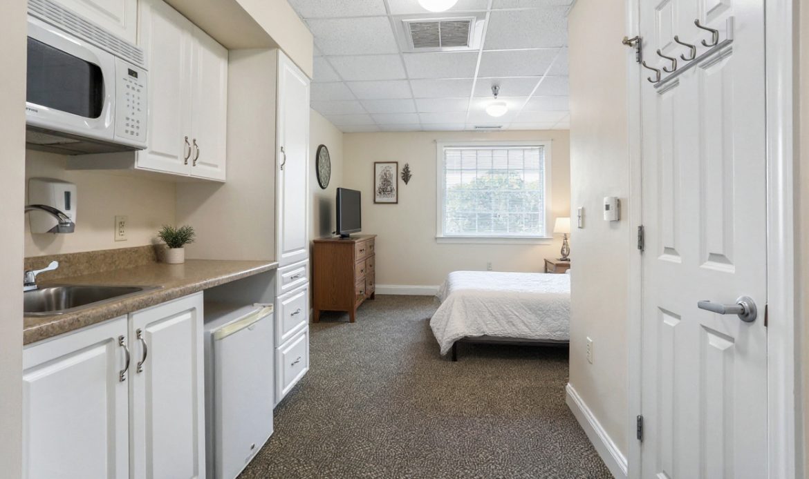 Compact room with kitchenette, bed, and TV. White cabinets, small fridge, microwave, and window illuminate beige space. Cozy, simple decor with wall hangings.