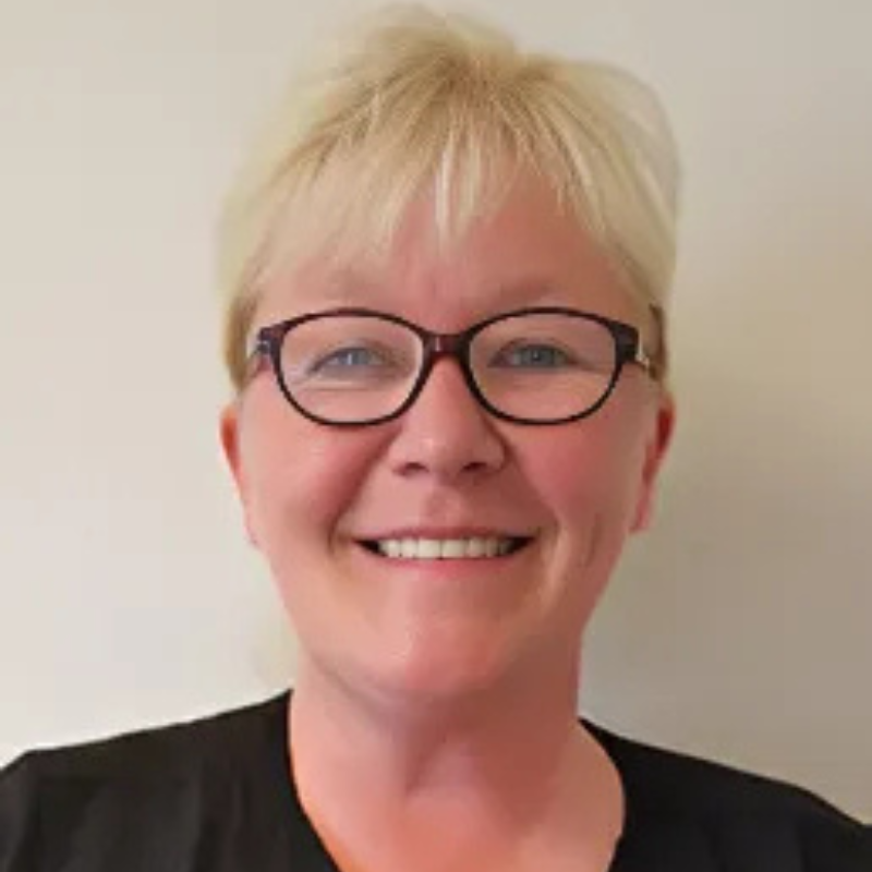 A smiling person with short blonde hair and glasses, wearing a black top, against a plain, light-colored background.