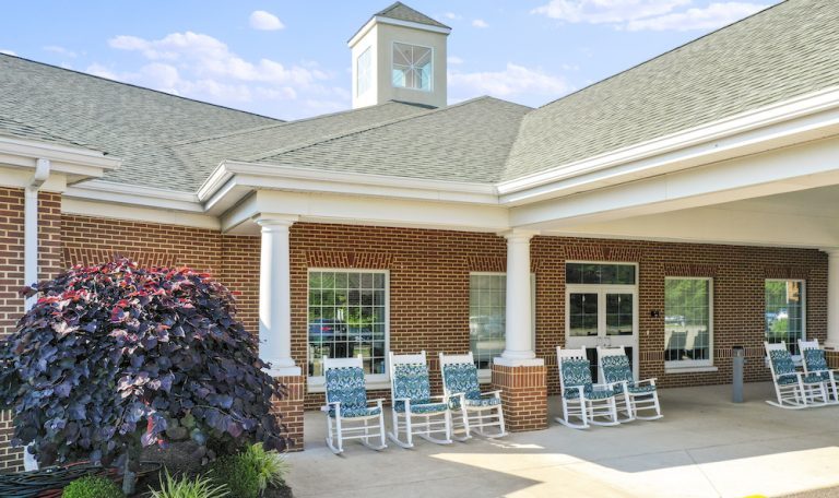 A brick building with white columns and rocking chairs on the porch, surrounded by neatly landscaped greenery under a clear blue sky.