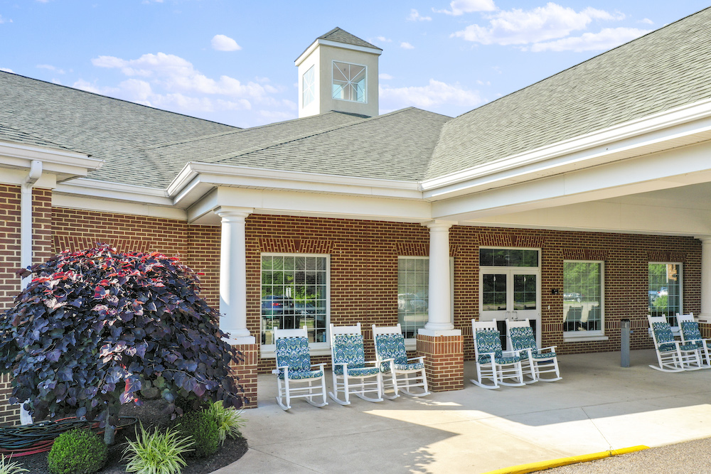 A brick building with white columns and rocking chairs on the porch, surrounded by neatly landscaped greenery under a clear blue sky.