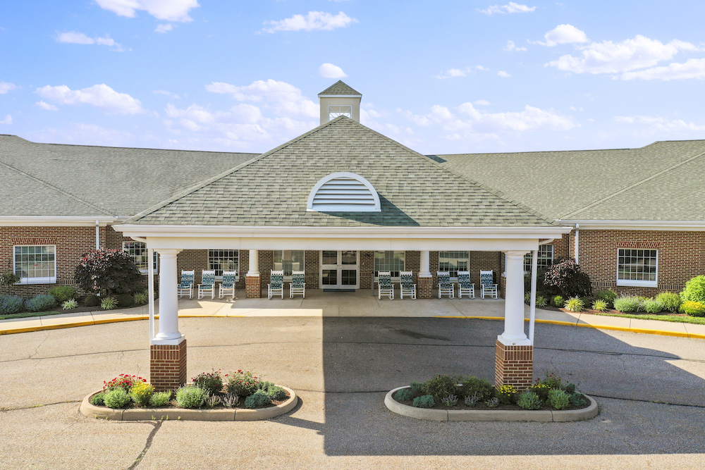 A brick building with a covered entrance features white pillars, rocking chairs, and surrounding greenery under a blue sky with scattered clouds.