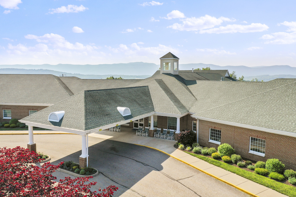 A large brick building with a covered entrance, landscaped gardens, and mountainous background under a clear blue sky. No recognizable landmarks visible.