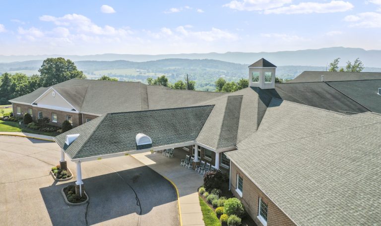 Aerial view of a sprawling building with a covered entrance. Surrounding lush landscape and distant mountains create a serene backdrop.