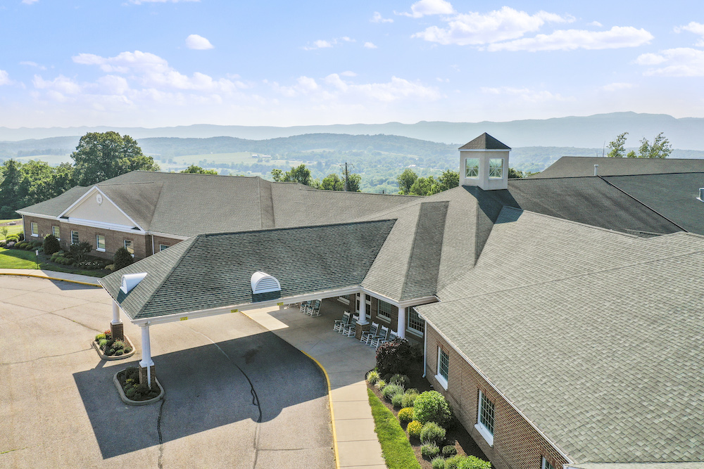 Aerial view of a sprawling building with a covered entrance. Surrounding lush landscape and distant mountains create a serene backdrop.