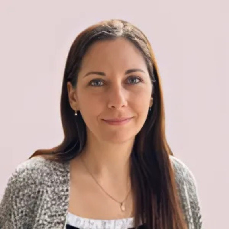 A person with long dark hair wearing a gray sweater and earrings, set against a plain light-colored background.