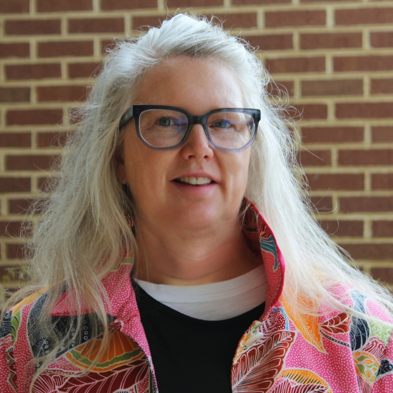 A person with glasses and long hair stands against a brick wall, wearing a colorful patterned shirt and a slight smile.