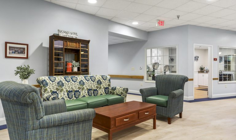 Room with floral-patterned sofa, armchairs, wooden table, US Post Office cabinet, and potted plants. Bright lighting and neutral-toned walls.