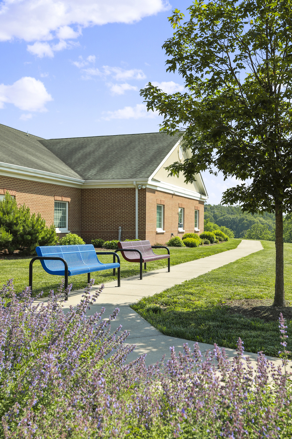 A brick building with a gable roof is surrounded by greenery, benches, and a walkway under a clear sky.
