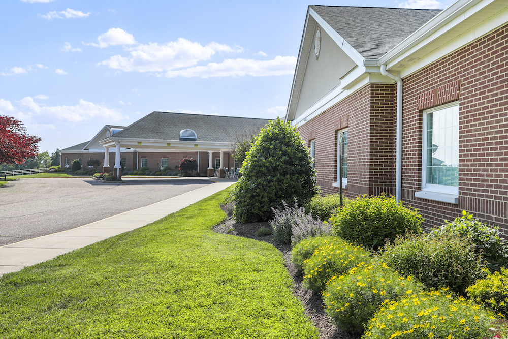 Hidden Springs Senior Living Assisted, Advanced, and memory care. Red-brick building with manicured landscaping under a blue sky. No people are visible. It&#039;s a suburban setting with neatly maintained surroundings.