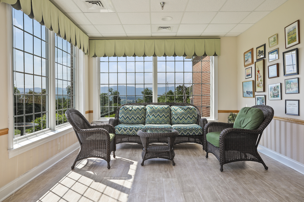 Assisted Living 29 A cozy sunroom with wicker furniture, patterned cushions, and wall art, offering scenic views through large windows with lush greenery outside.