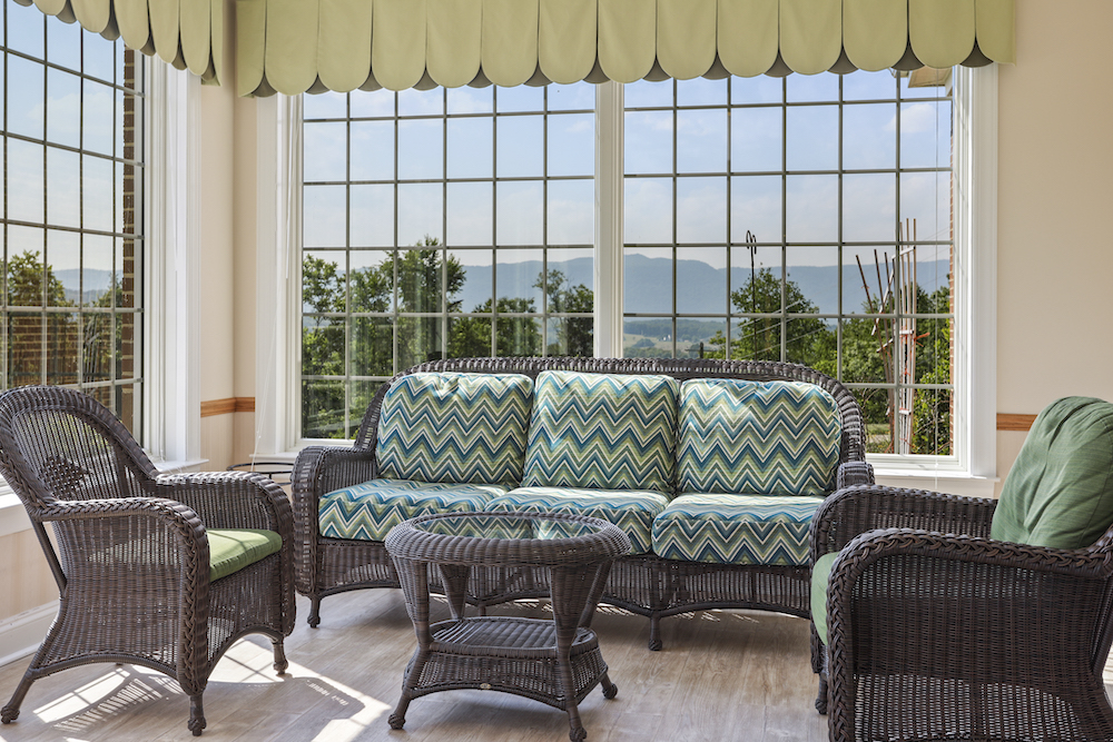 Assisted Living 4 Sunlit room with wicker furniture, green cushions, large windows, and mountain views. The serene setting creates a cozy, inviting atmosphere for relaxation.