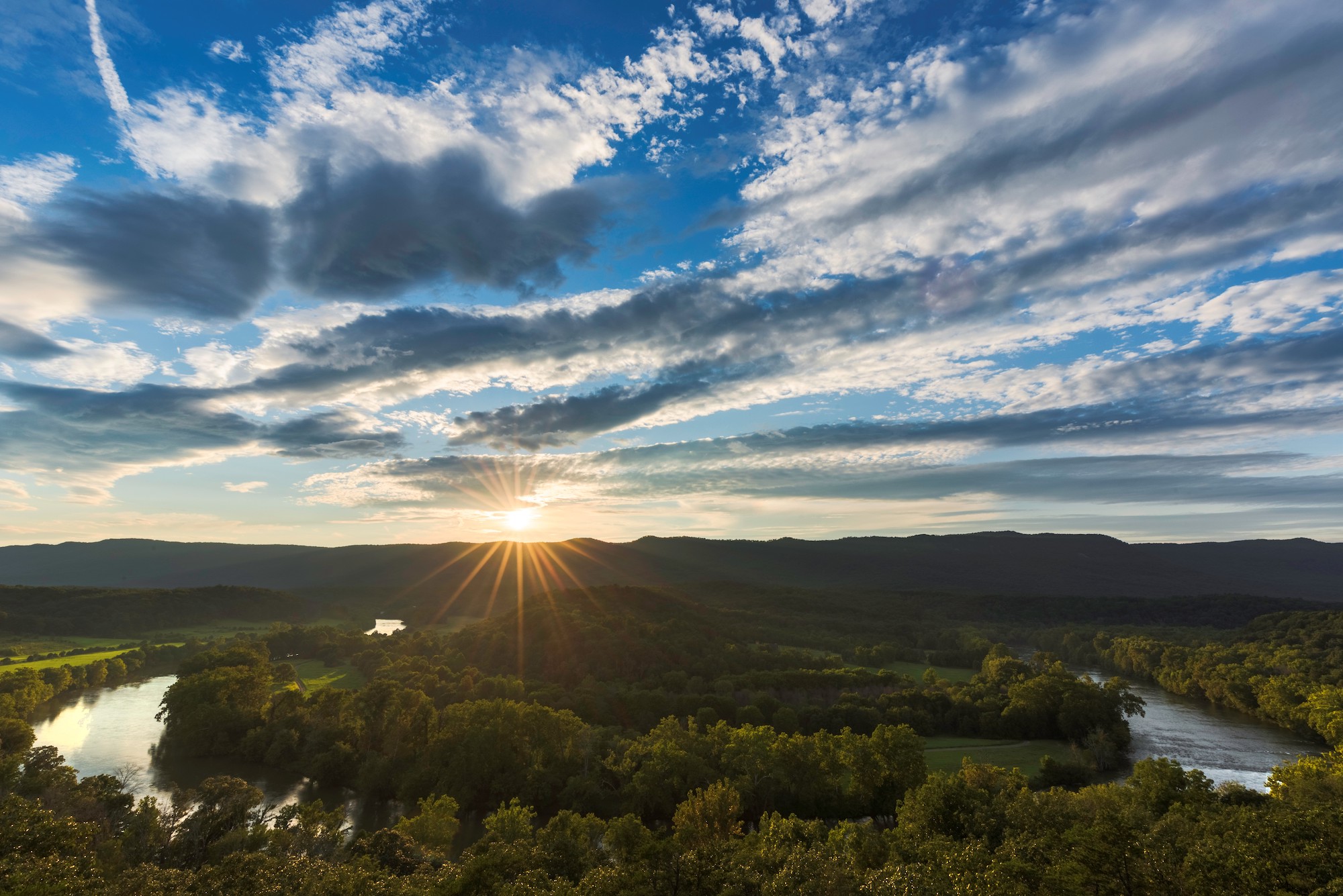 Sunset over a lush, forested landscape with a winding river beneath a dramatic, cloud-filled sky. No recognizable landmarks or historical buildings visible.