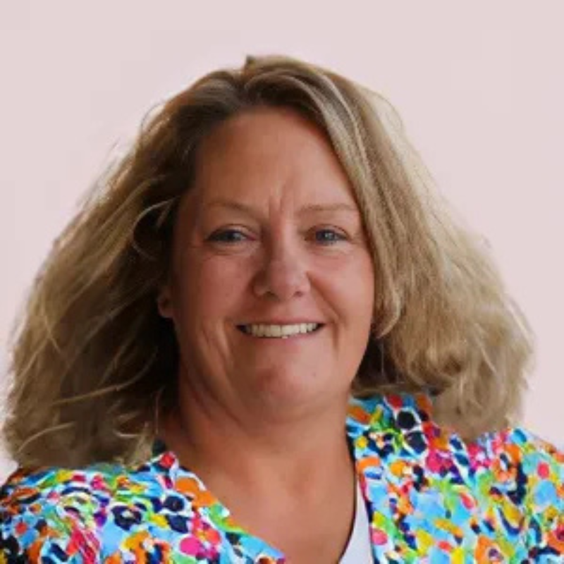 A person with shoulder-length hair smiles, wearing a colorful patterned top against a plain background. No landmarks or historical buildings are visible.