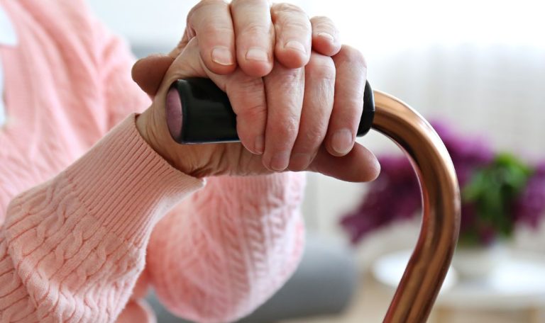 A person with folded hands rests on a cane. They wear a pink sweater. Blurred lilacs in the background suggest a cozy setting.