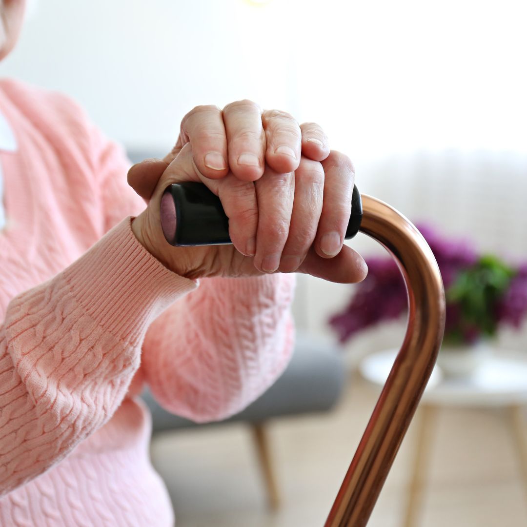 A person with folded hands rests on a cane. They wear a pink sweater. Blurred lilacs in the background suggest a cozy setting.