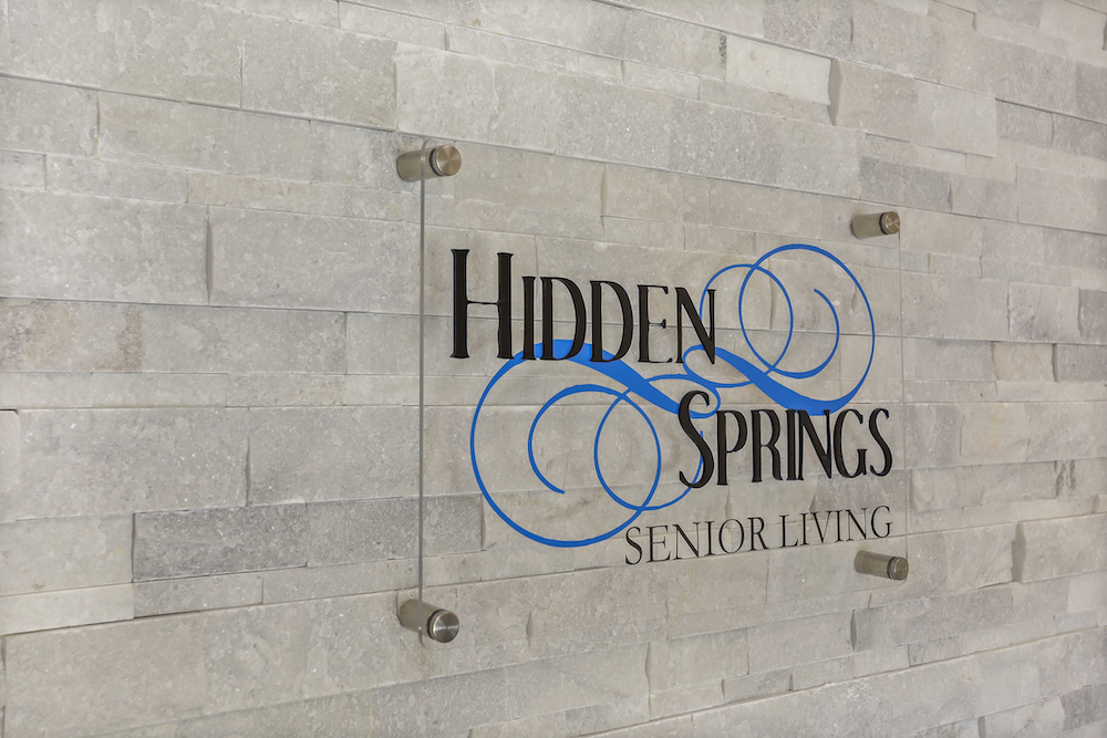 The image shows a glass sign reading &quot;Hidden Springs Senior Living&quot; mounted on a light stone wall with decorative blue swirls.