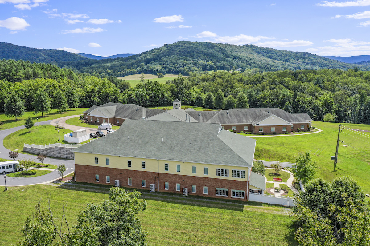 A large building complex surrounded by lush green hills and trees under a clear blue sky, with a paved pathway and parked vehicles.