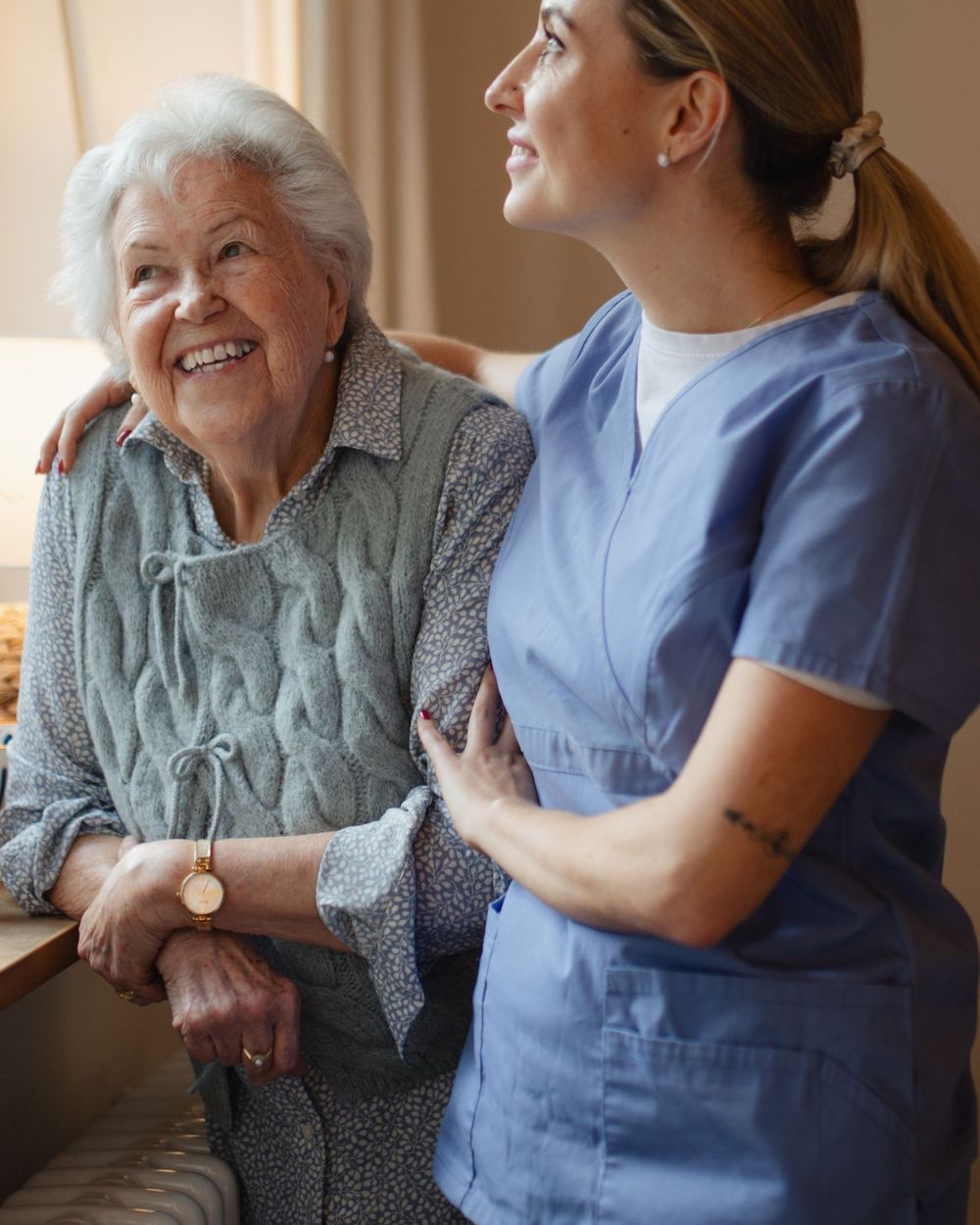 Two people smiling together; one in a blue uniform. They appear content indoors, suggesting companionship and care in a warm, bright setting.
