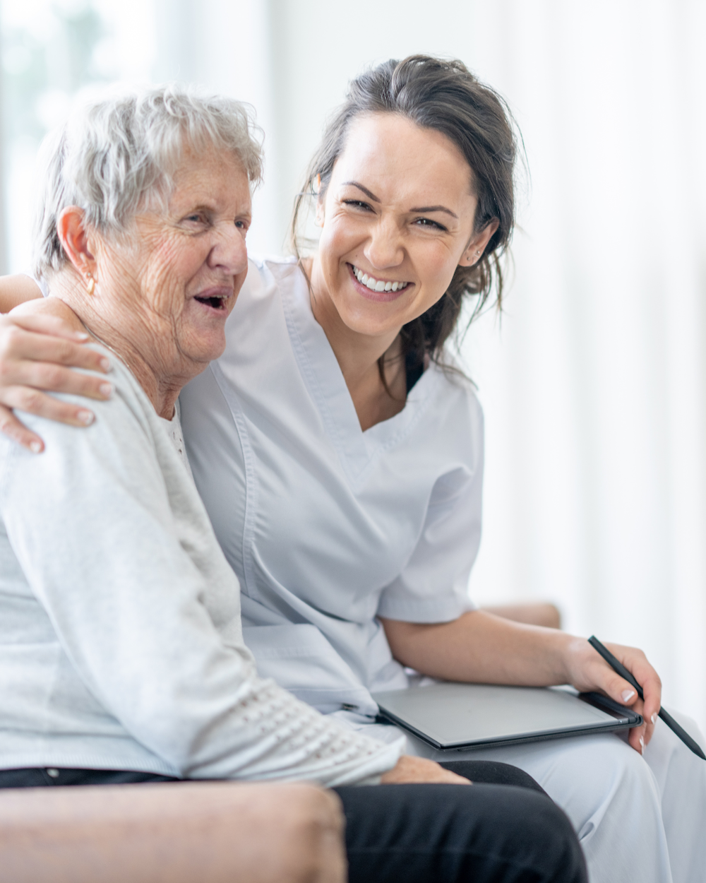 A person in a white uniform smiles while embracing an older person, creating a warm, supportive atmosphere in a well-lit room.