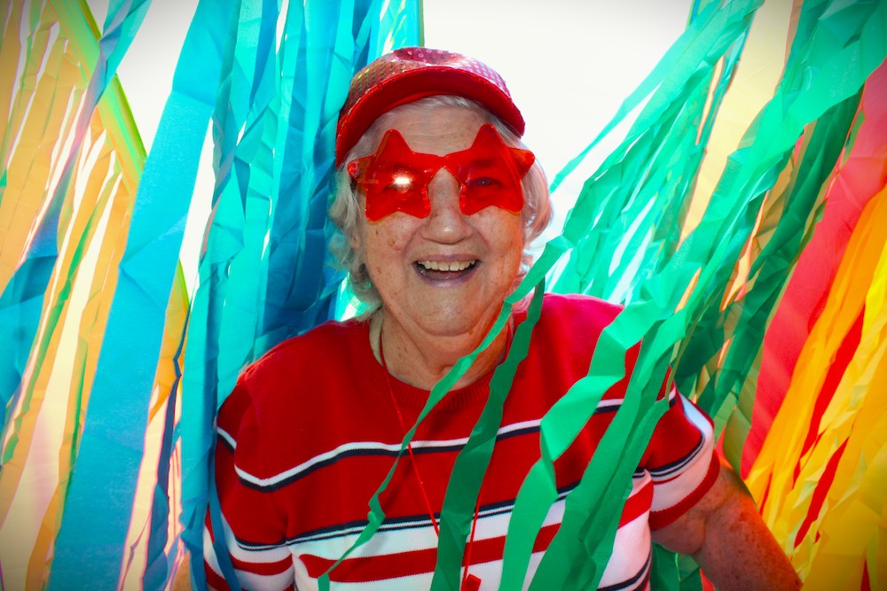 A person wearing star-shaped glasses and a cap smiles amidst vibrant, colorful streamers in a festive atmosphere.