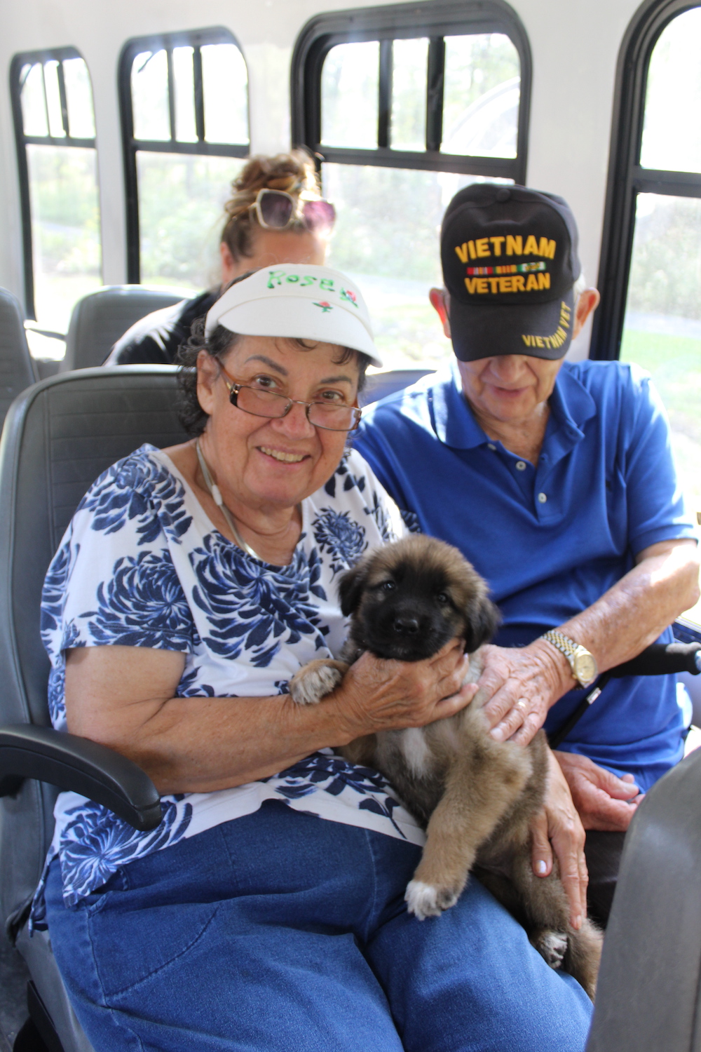 Two people sit in a vehicle holding a small puppy. One wears a &quot;Vietnam Veteran&quot; cap. Another person is seated behind.