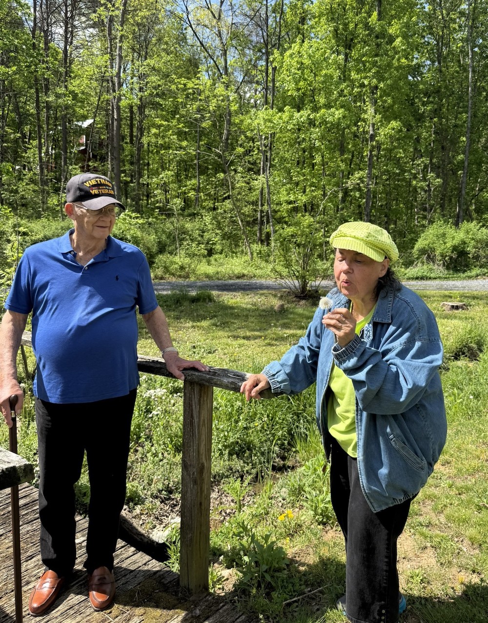 Two people outdoors enjoy a sunny day in a lush, green forest setting. One person holds a dandelion.