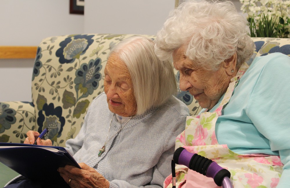 Two elderly people sit together on a floral couch, engrossed in filling out a form or activity using a pen at a community living facility.