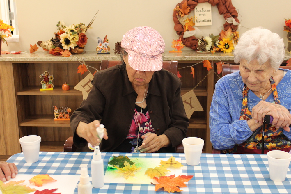 Two people doing fall-themed crafts at a table with leaves and decorations, surrounded by autumn decor on shelves and a wall wreath.
