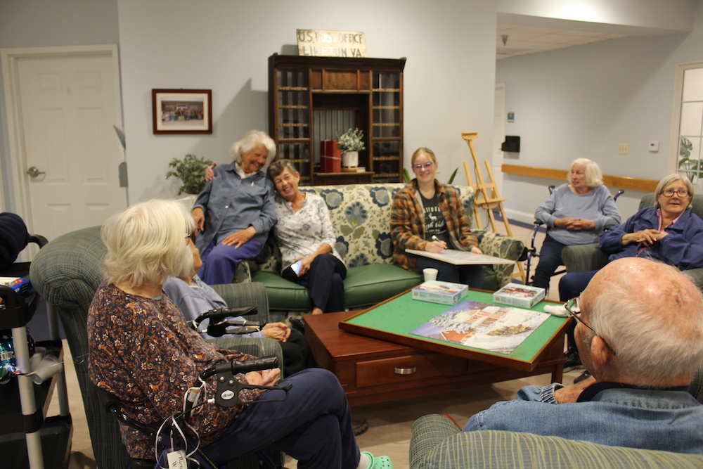 A group of eight people sit together in a cozy room, surrounded by puzzles and comfortable seating, enjoying a warm gathering.