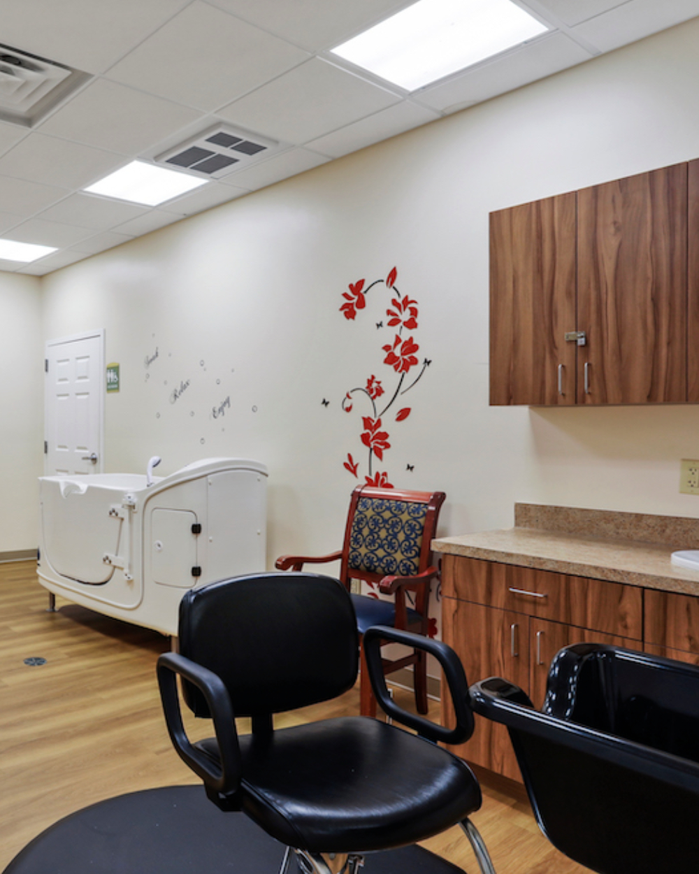 A salon interior with two chairs, floral wall art, a sink, and wooden cabinets. Room features a bathing tub and wooden flooring.