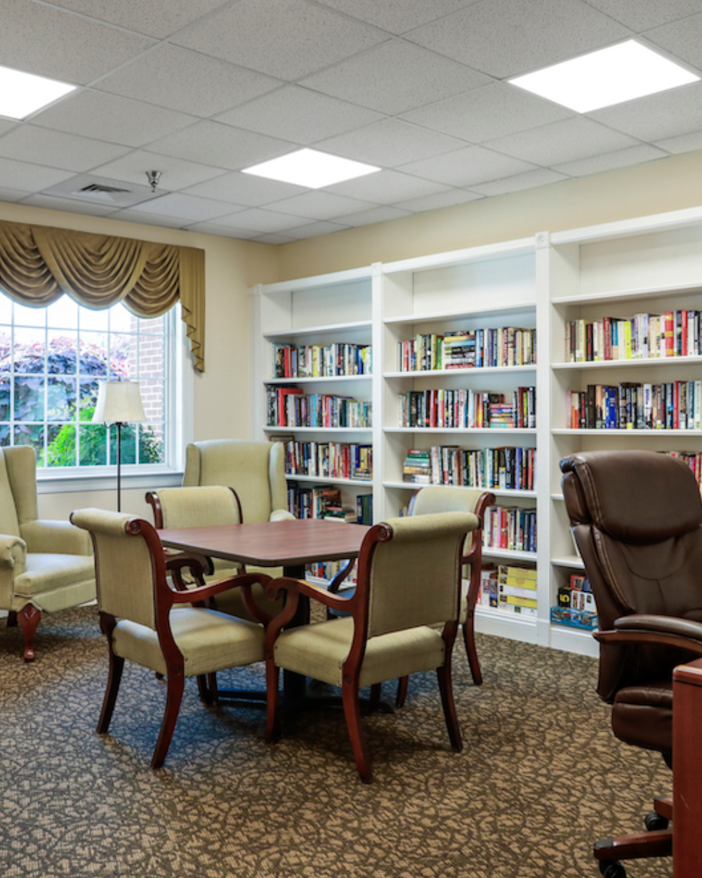 Cozy library room with bookshelves, a table, and armchairs. Large window with drapes provides natural light. Brown carpet complements the setting.