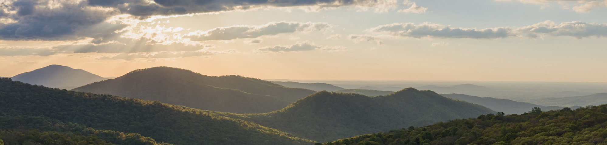 Rolling Blue Ridge Mountains under a cloudy sky, with sunlight filtering through, creating a serene landscape. Lush green hills spread across the horizon.