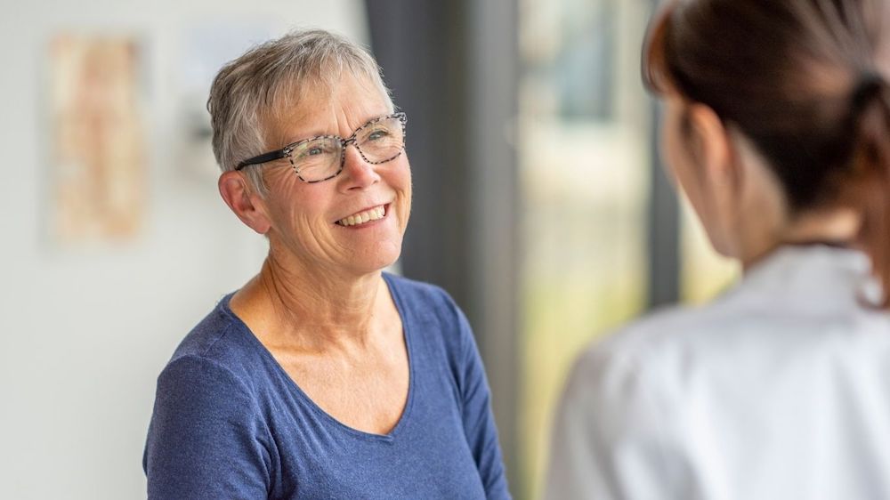 Two people engaged in a friendly conversation indoors, one smiling warmly. Neutral background and soft lighting create an inviting, casual atmosphere.