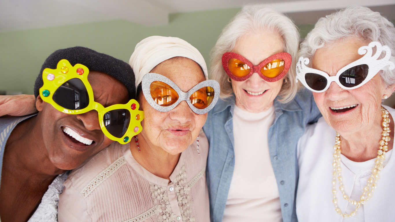 Four people smiling, wearing colorful, oversized sunglasses, standing closely indoors, creating a joyful and playful atmosphere. No landmarks or historical buildings visible.