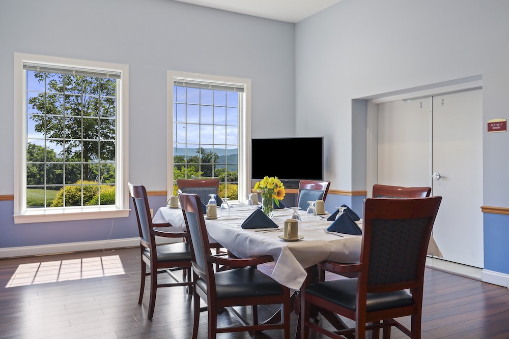 Assisted Living 2 A well-lit dining room with neatly set table, flowers, and a scenic view through large windows. There's a television and labeled door.