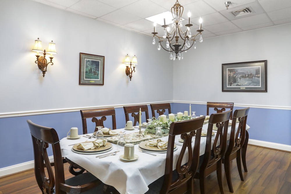 Assisted Living 23 Elegant dining room with chandelier, wooden chairs, and a table set for eight. Wall features paintings, sconce lighting, and two-tone decor.