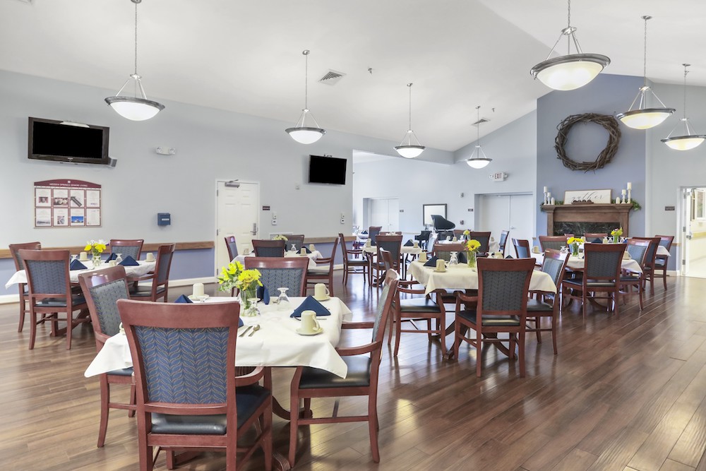 Assisted Living 26 Bright dining room with wooden floor, neatly arranged tables, and decorative lighting. Flowers and napkins accent tables. TVs are mounted on the walls.