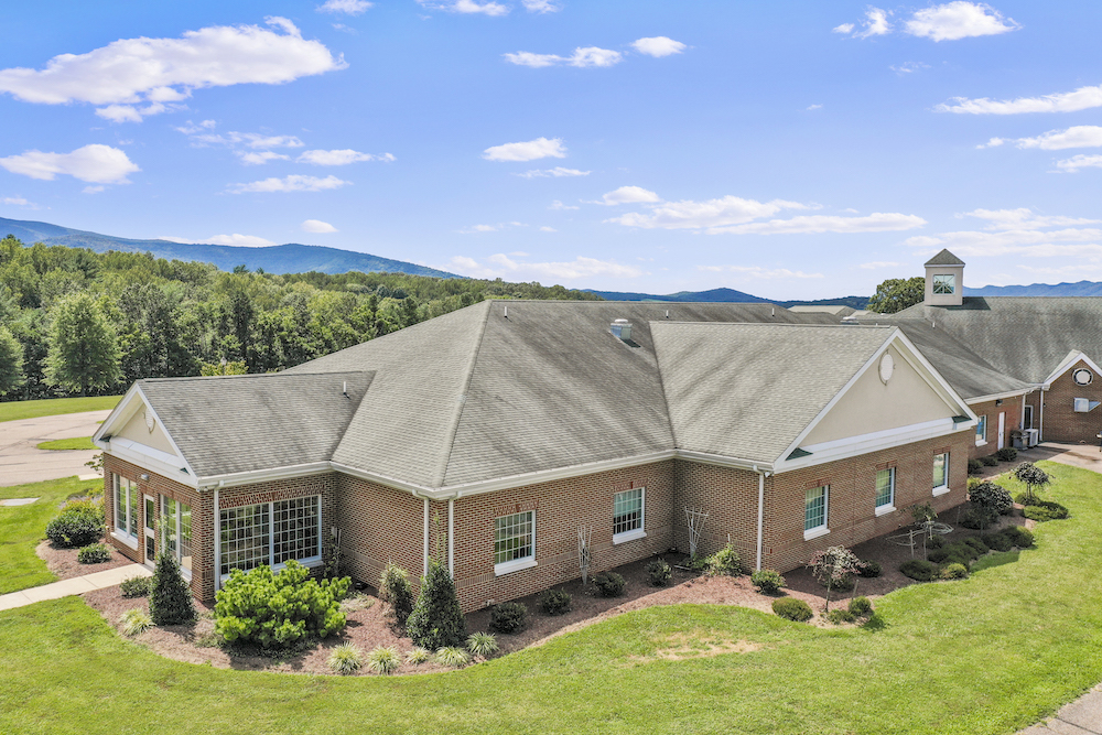 Assisted Living 39 Brick building with gabled roof, surrounded by green lawns and trees. Hills and blue sky with clouds in the background.