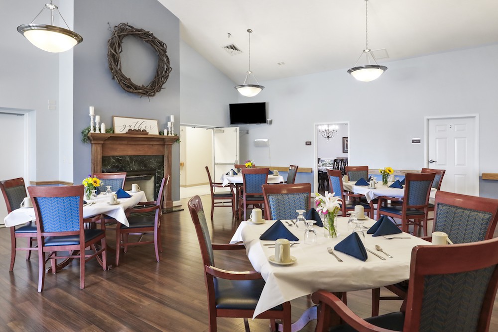 Assisted Living 21 Cozy dining area with wooden tables, chairs, and floral centerpieces. A decorative wreath above a fireplace creates a welcoming atmosphere.