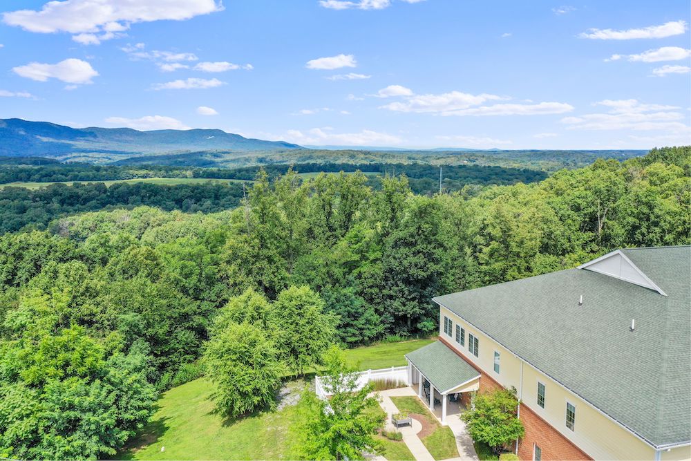 Assisted Living 35 Large building beside lush forest, with mountain range in the background under a clear blue sky. Pathway leads to building entrance.