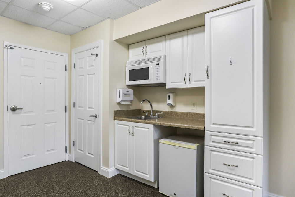 Assisted Living 22 Compact kitchen with white cabinets, microwave, mini-fridge, and sink. Beige walls and carpeted floor. Doors on the left side.