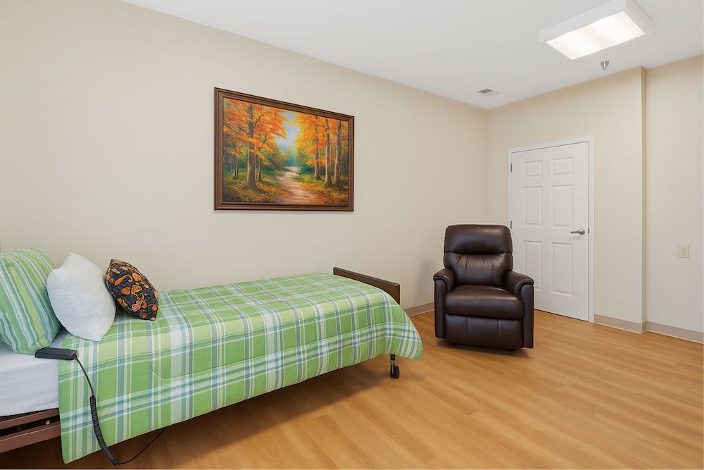 A tidy room with a bed, green plaid bedding, brown recliner, and a forest painting on the wall. No people present.