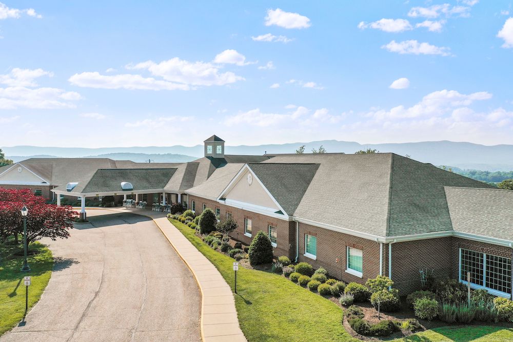 Assisted Living 31 A large brick building with a driveway and manicured gardens, set against a backdrop of distant mountains under a partly cloudy sky.
