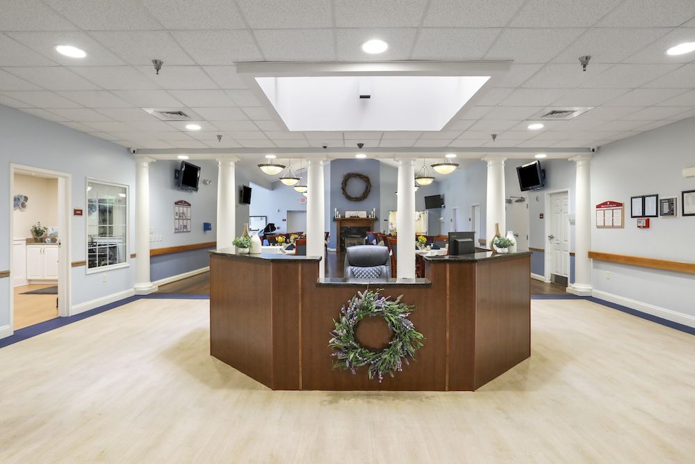 Assisted Living 16 A reception area with wooden desk, decorative wreath, columns, and plants. Soft lighting enhances the cozy environment. Multiple doors and wall-mounted screens visible.