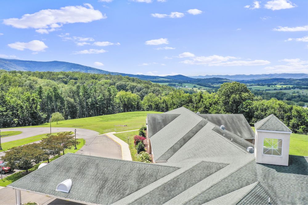 Assisted Living 14 Aerial view of a large building with a green roof, surrounded by lush greenery and distant mountains under a clear blue sky.