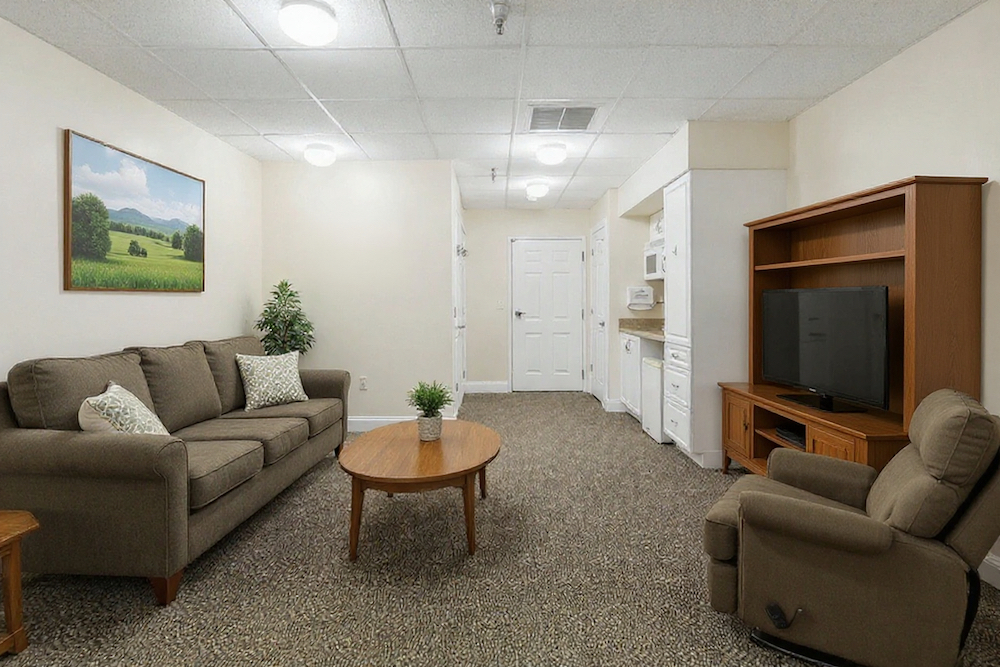 Assisted Living 12 Cozy living room with beige sofa, recliner, TV on a wooden stand, and a landscape painting. Door and kitchenette in the background.