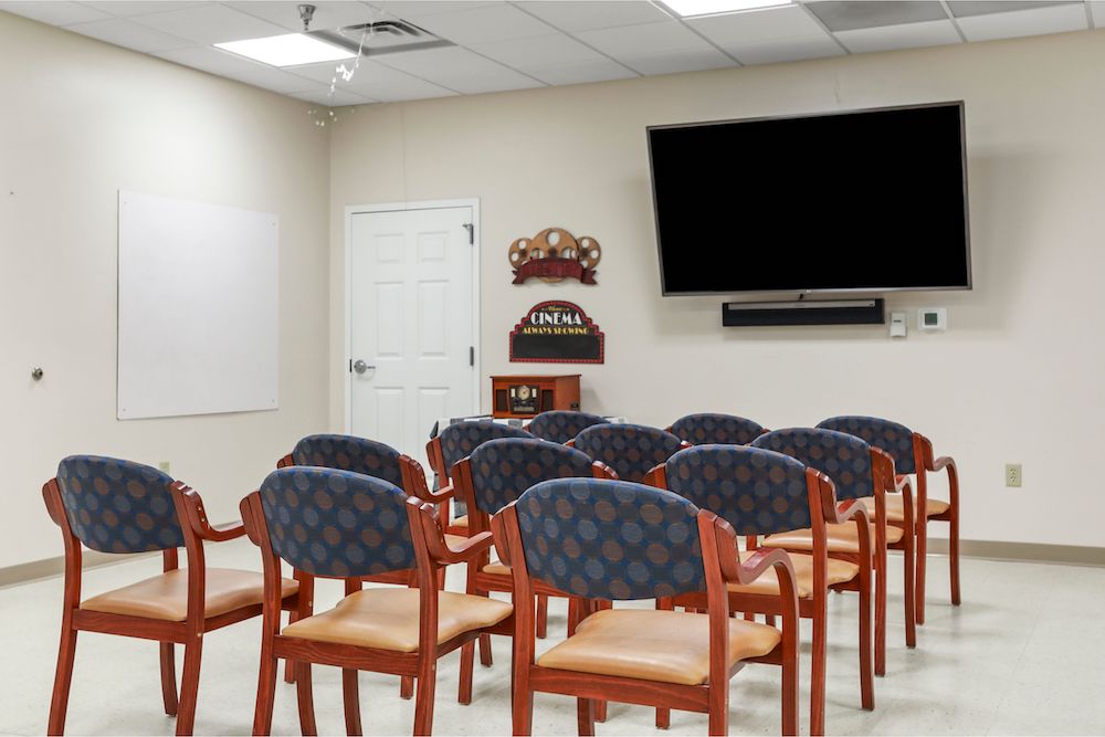 Assisted Living 8 A small room with rows of chairs faces a large screen, a "Cinema" sign, and enclosed door on the wall.