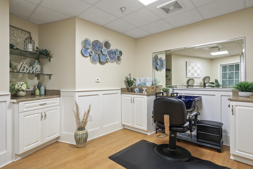 Assisted Living 6 Hair salon room with modern decor, featuring a black chair, large mirror, decorative wall art, and white cabinets on light wood flooring.